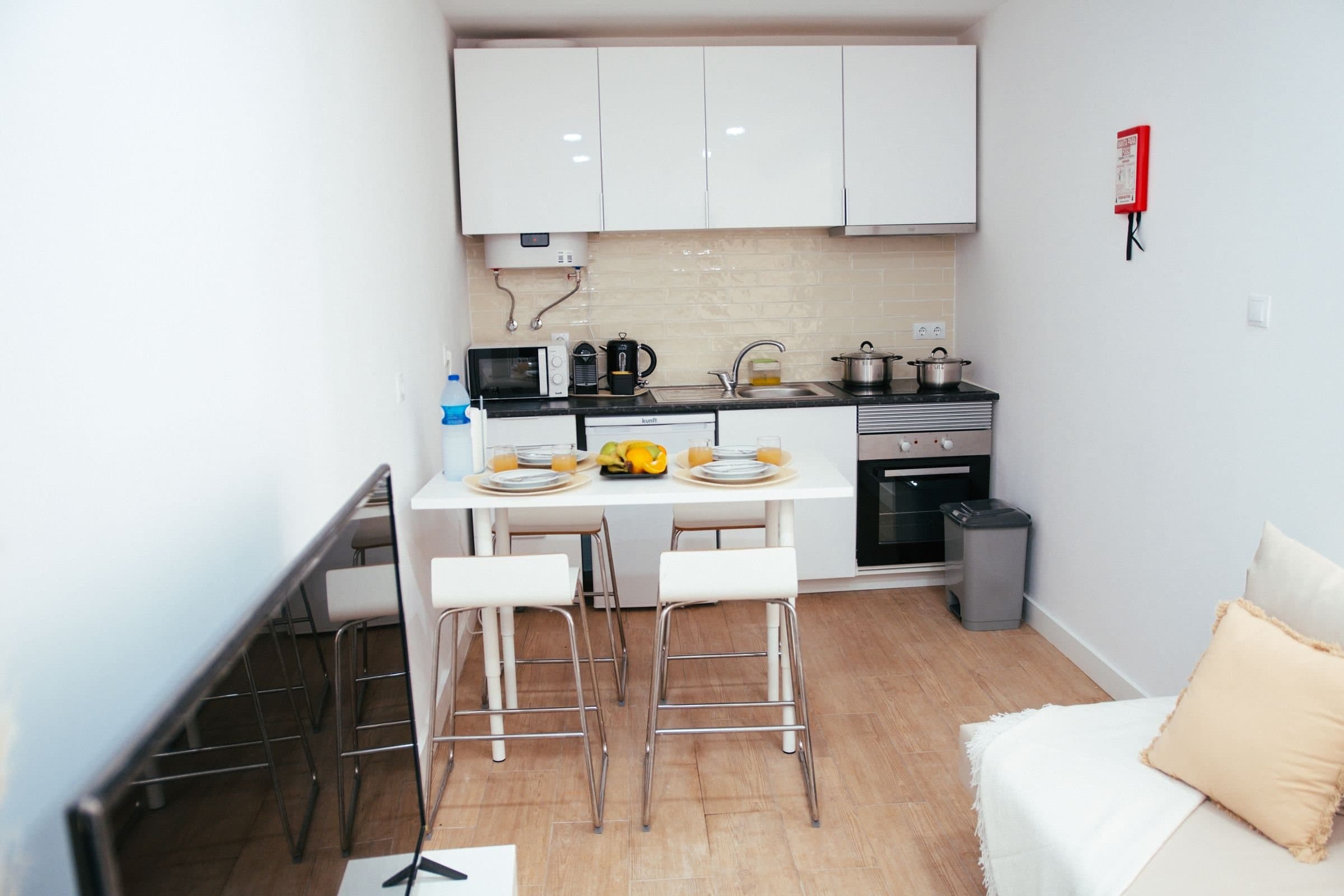 Compact flat kitchenette with a wall-mounted water heater, microwave, kettle, oven and hob, and a white breakfast bar with stools