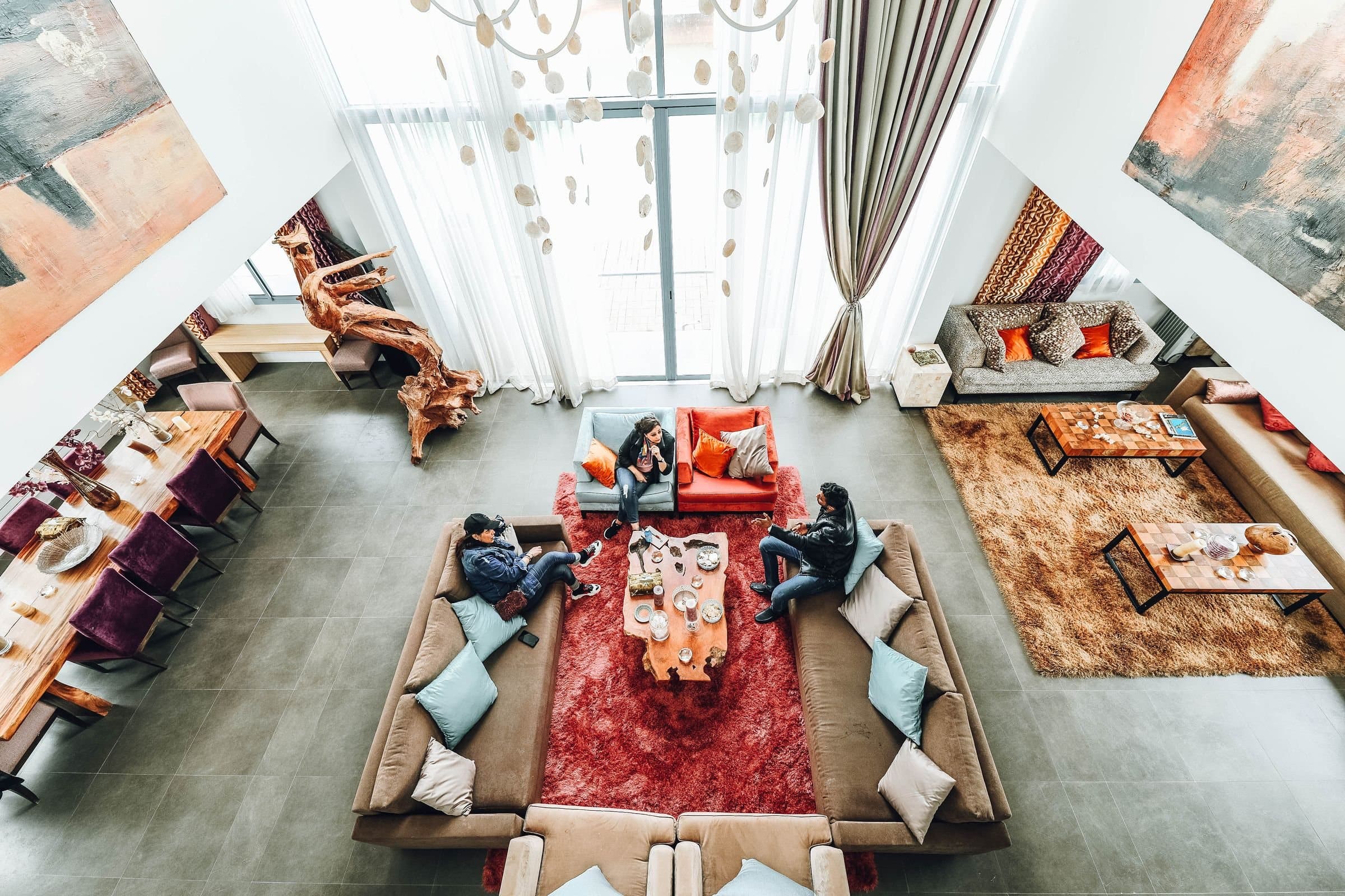 Three people chat on facing sofas in a high-ceilinged shared lounge, seen from above, beside a long communal dining table