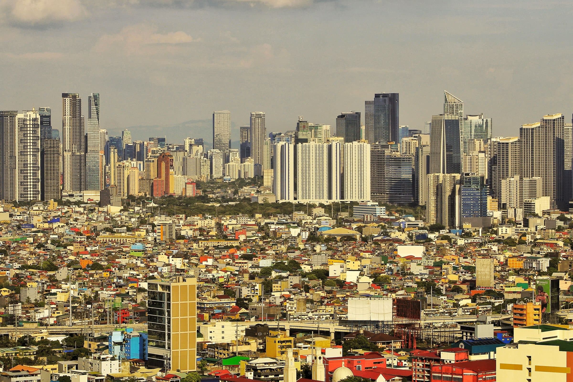 Manila's high-rise towers rising behind a dense spread of low-rise homes and colourful rooftops under a hazy sky