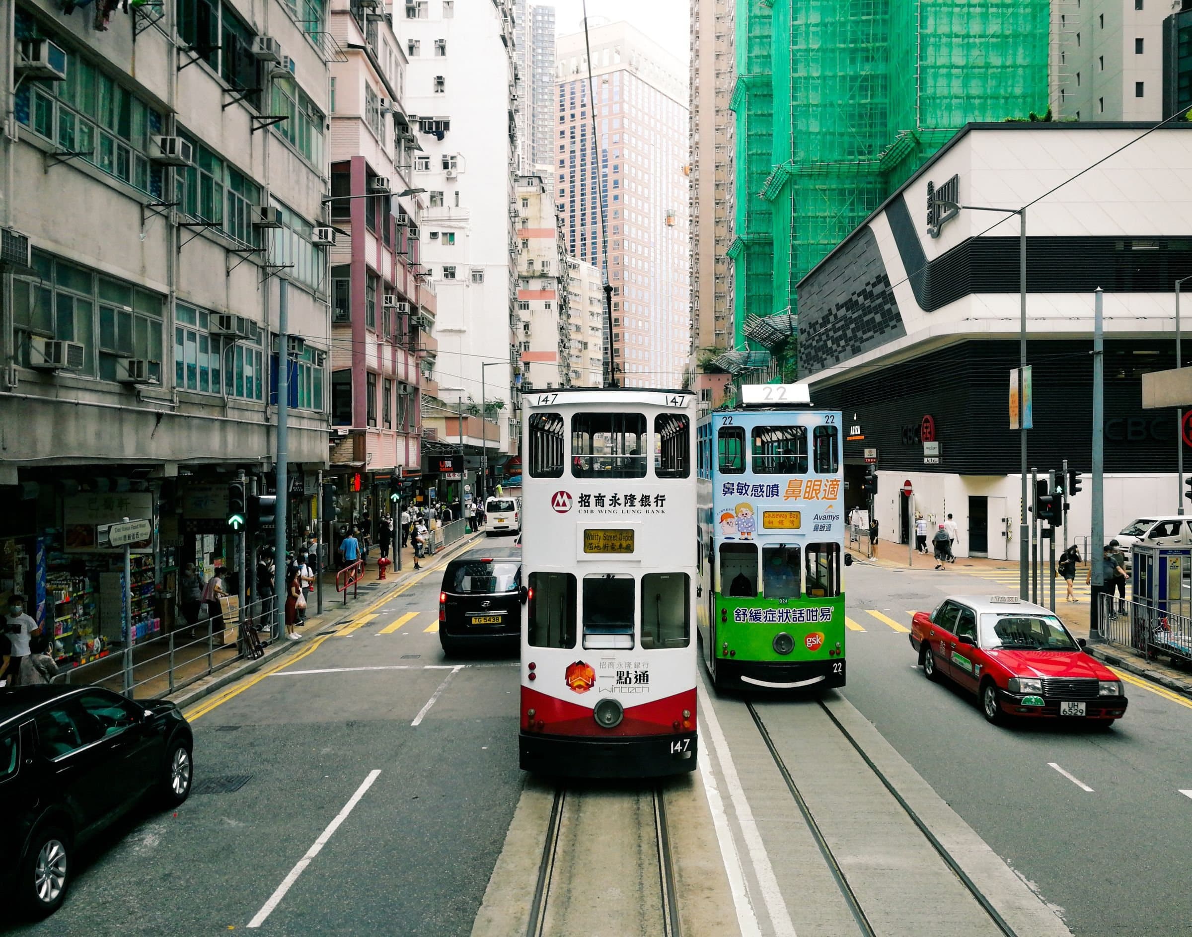Two double-decker trams and a red taxi on a busy Hong Kong Island street lined with shops and apartment blocks
