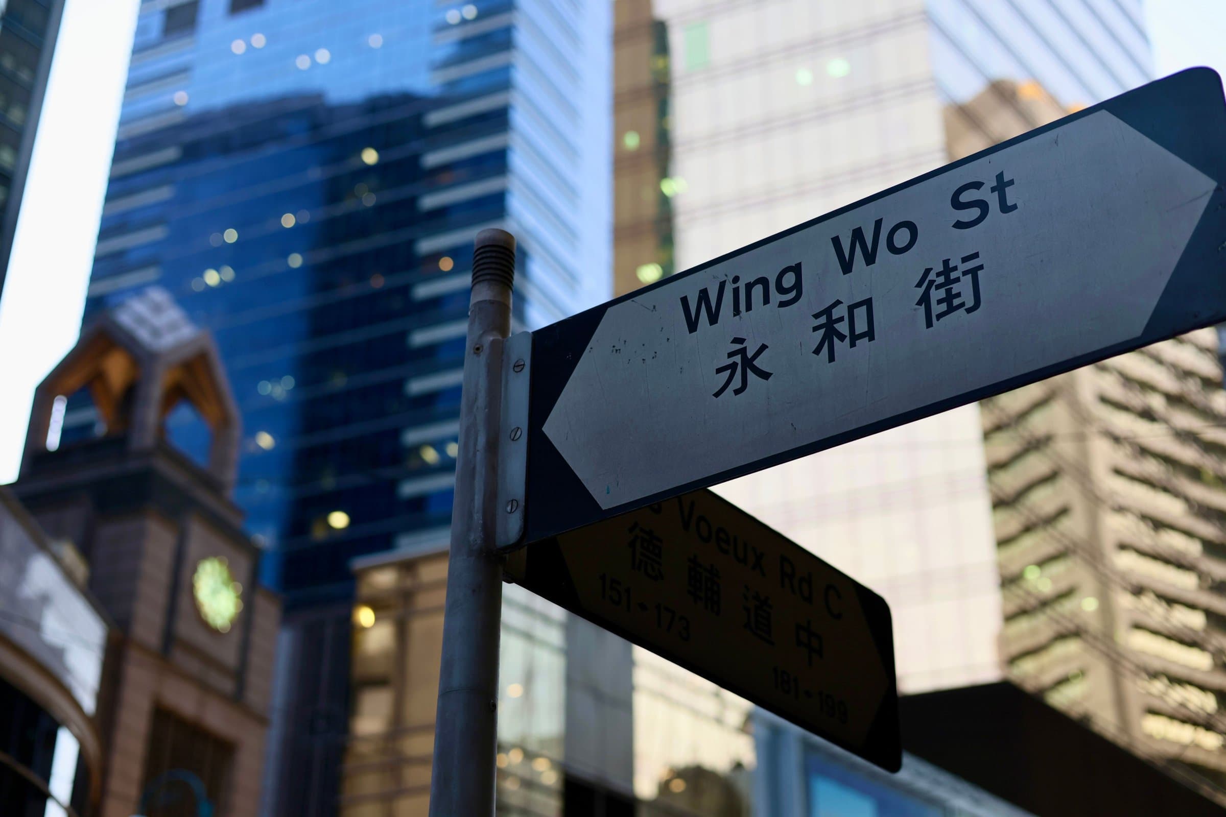 Bilingual street signs in Sheung Wan, Hong Kong: Wing Wo St 永和街 above Des Voeux Rd C 德輔道中 with house numbers 151–173 and 181–199, glass towers behind